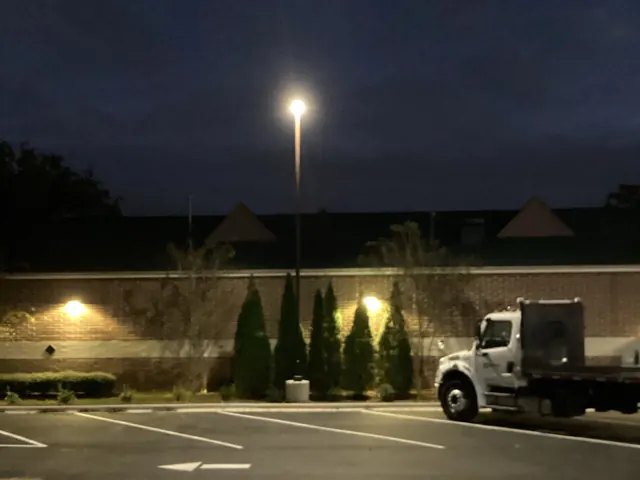 Parking lot at night lit by a tall pole light and two wall-pack lights on a brick building, with a white work truck parked at the right