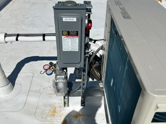 Gray rooftop disconnect switch on a metal strut frame beside an HVAC condensing unit, fed by liquid-tight flexible conduit