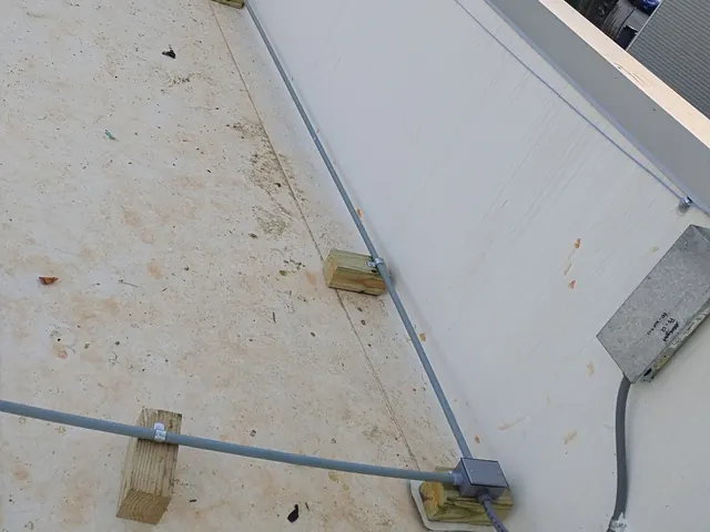 Gray conduit runs supported on wood blocks across a flat white roof to a junction box and a wall-mounted pull box