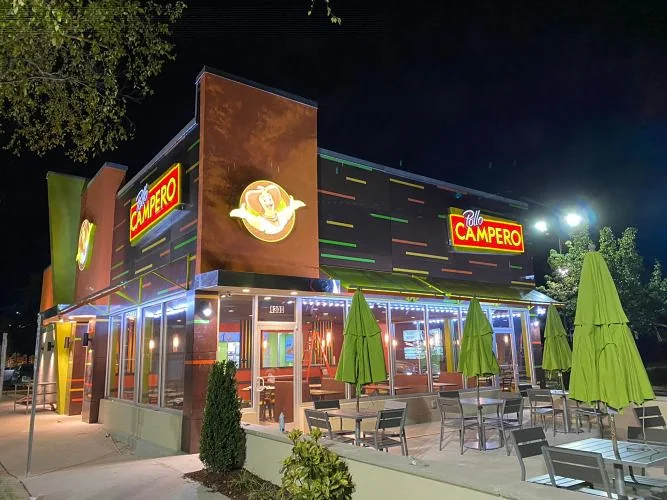 Pollo Campero restaurant in Raleigh, NC at night, with illuminated Pollo Campero signs, a lit glass storefront and a patio with closed green umbrellas